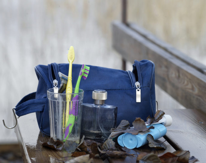 Toothbrushes in the drinking glass next to body lotion and neseser on the wet bench, outdoor shot, concept of travel