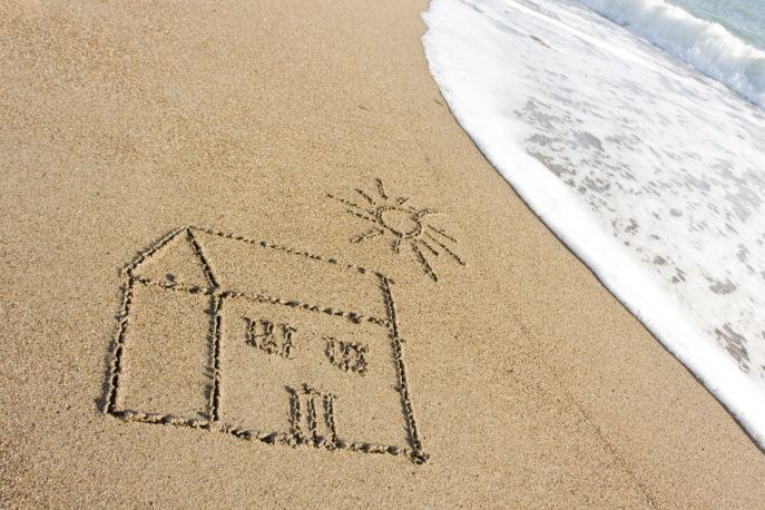 photo of house drawing on beach sands by mediterranean sea.There is a drawing of sun on house which is positioned on left side of frame.The sea waves are on the right side.Focus is on house.A full frame DSLR camera was used to shot photo.